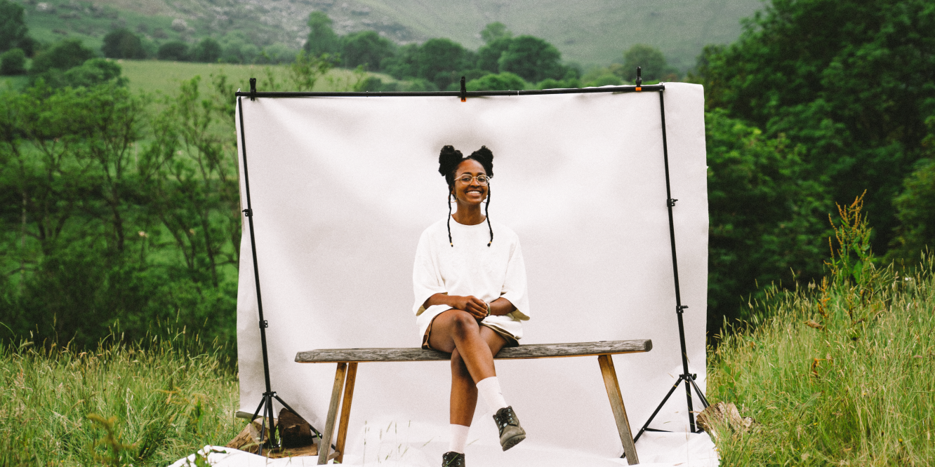 About - Emma Blake Morsi sat on a bench in front of a small white back-drop in the middle of a field with mountains in the far background