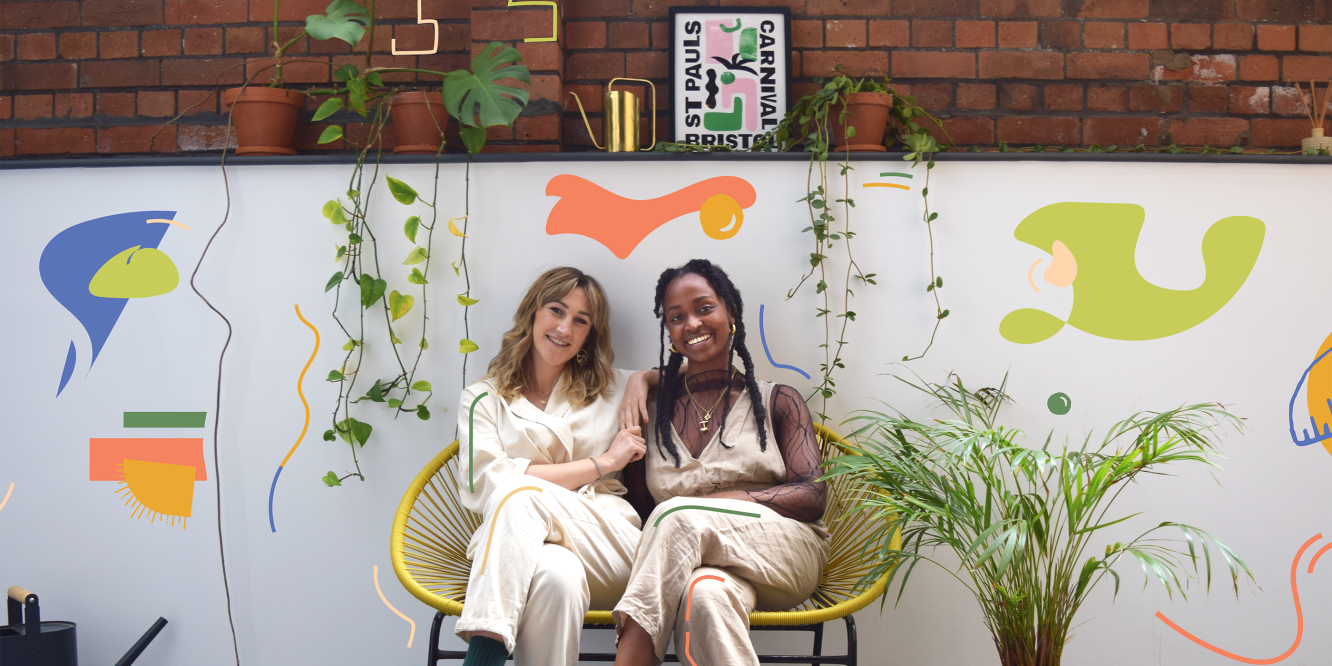 Katie, the founder, and Emma, the producer, sat together on a curved yellow bench. Although dressed similarly in beige jumpsuits, Katie is a mid 20s white woman with shoulder-length blonde hair and big, bright smile. Emma is a mid 20s dark-skinned Black woman, also wearing a long-sleeved mesh top and gold jewellery. Behind them is a short white wall, on top of which hang small, long plants with a swiss-cheese looking monsterra plant, gold watering can and small black, green and pink typography print of 'St Pauls Carnival' on the shelf with a red brick wall behind. Besides Emma is a palm tree that is half the height of the white wall behind.