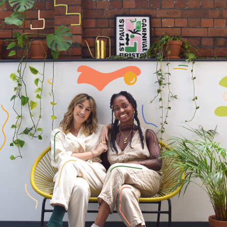Katie, the founder, and Emma, the producer, sat together on a curved yellow bench. Although dressed similarly in beige jumpsuits, Katie is a mid 20s white woman with shoulder-length blonde hair and big, bright smile. Emma is a mid 20s dark-skinned Black woman, also wearing a long-sleeved mesh top and gold jewellery. Behind them is a short white wall, on top of which hang small, long plants with a swiss-cheese looking monsterra plant, gold watering can and small black, green and pink typography print of 'St Pauls Carnival' on the shelf with a red brick wall behind. Besides Emma is a palm tree that is half the height of the white wall behind.